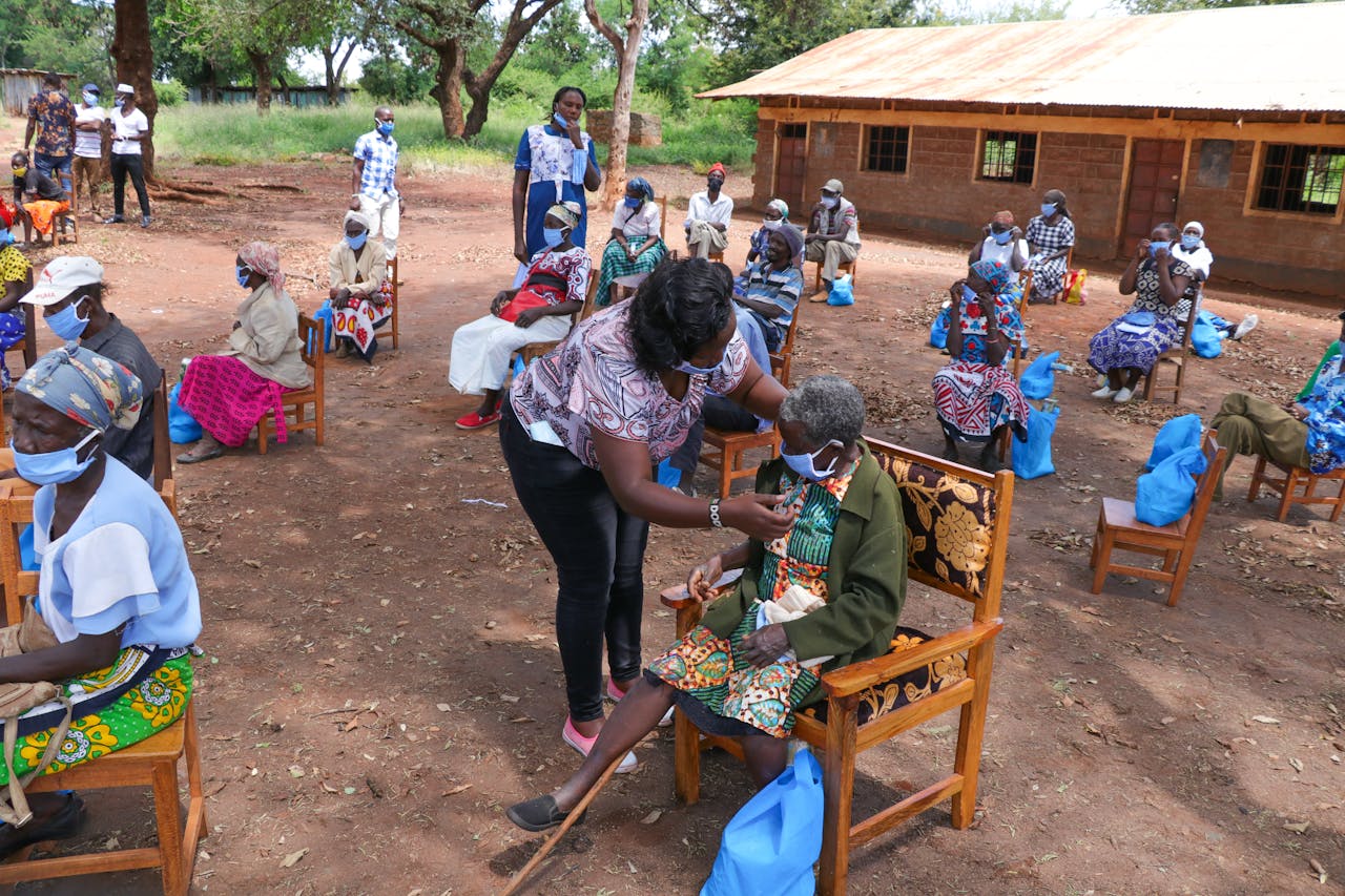 High angle of anonymous African female taking care of senior people in face masks sitting on chair near shabby building of nursing home in village