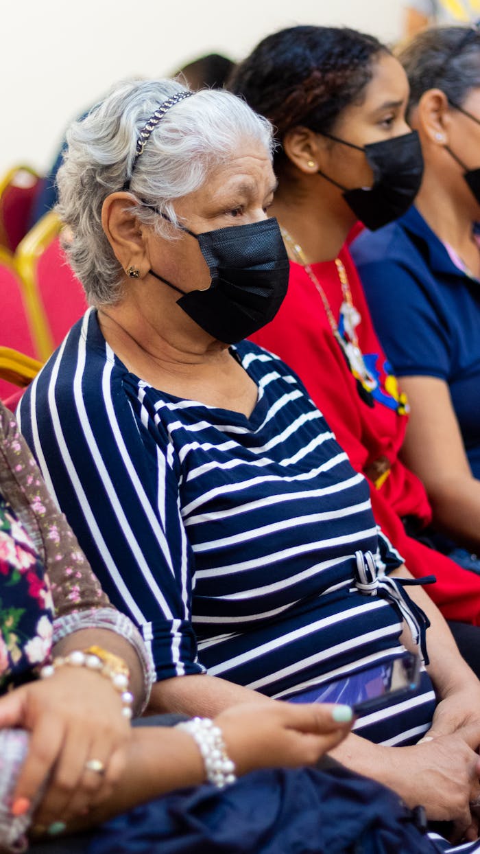 Elderly woman with gray hair and mask sitting with companions indoors.
